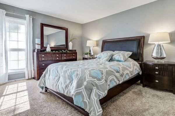 A modern bedroom with a wooden bed, patterned bedding, dresser with mirror, lamp, and light gray walls at Madison Hunters Glen Apartments.