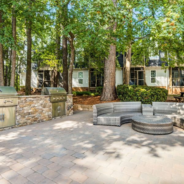 Outdoor patio with curved wicker seating, stone grill, and tall trees shading a modern house in the background at Madison Hunters Glen apartments.